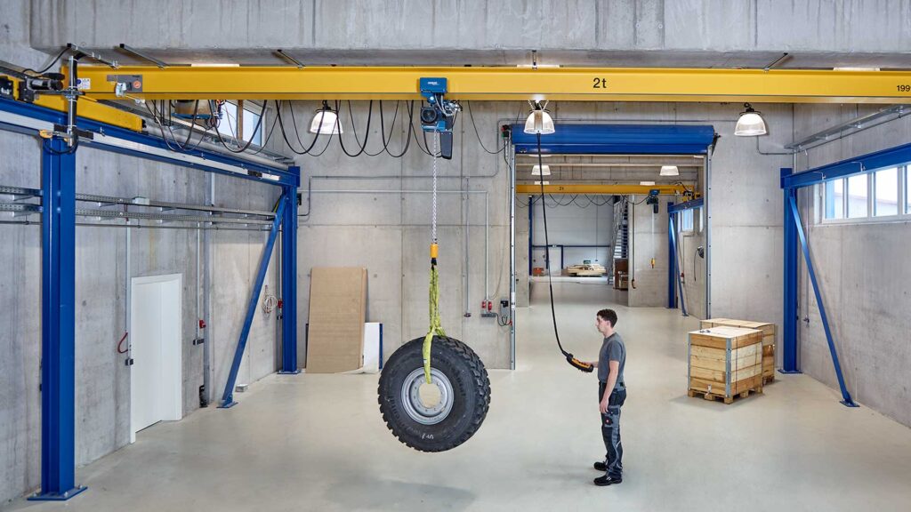 Demag DC Chain Hoist with E22C Trolley on a beam crane lifting a large vehicle tire. The operator is pressing the lift button on the pendant.