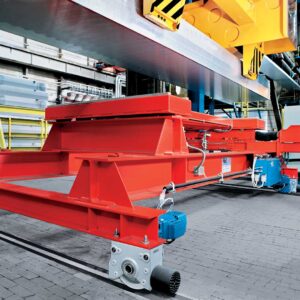 Red transfer car with Demag wheel blocks and motors sits as an overhead crane places a large piece of raw material for transport.