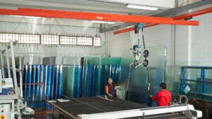 Demag KBK Crane and DC chain hoist with a pneumatic manipulator lifting a panel of glass. Two workers are handling the glass.